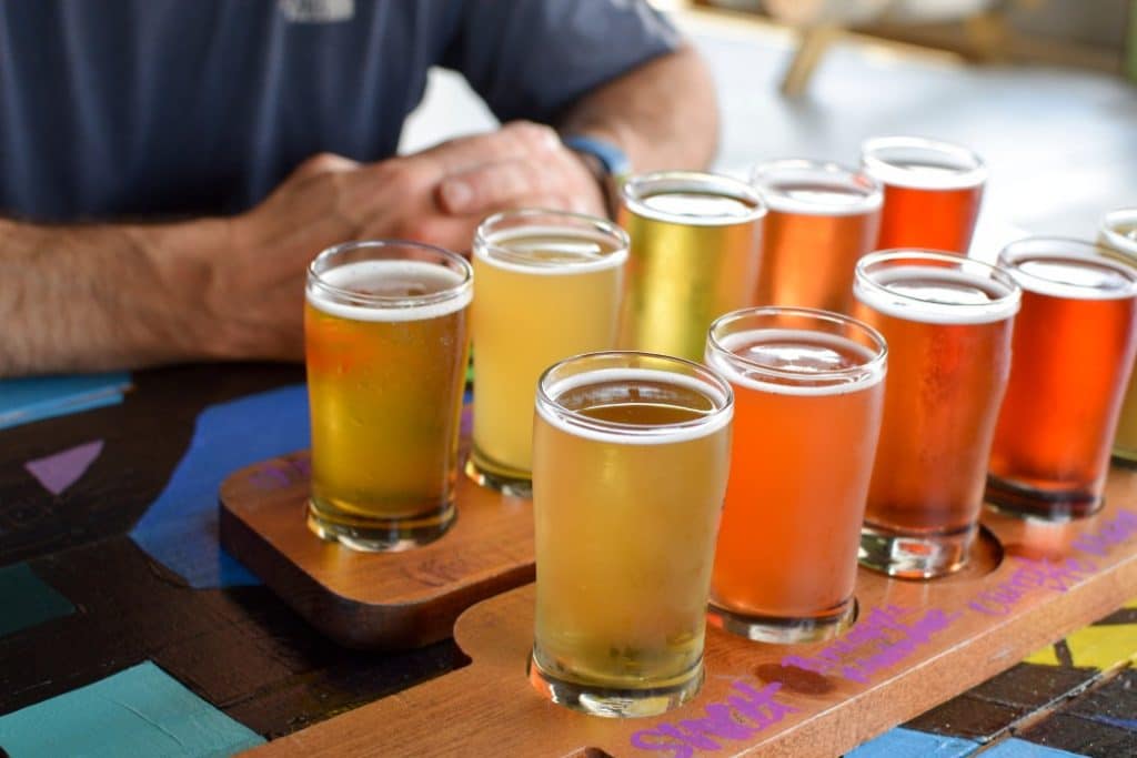 A flight of various craft beers is displayed on a wooden tray with a person's hands resting nearby.