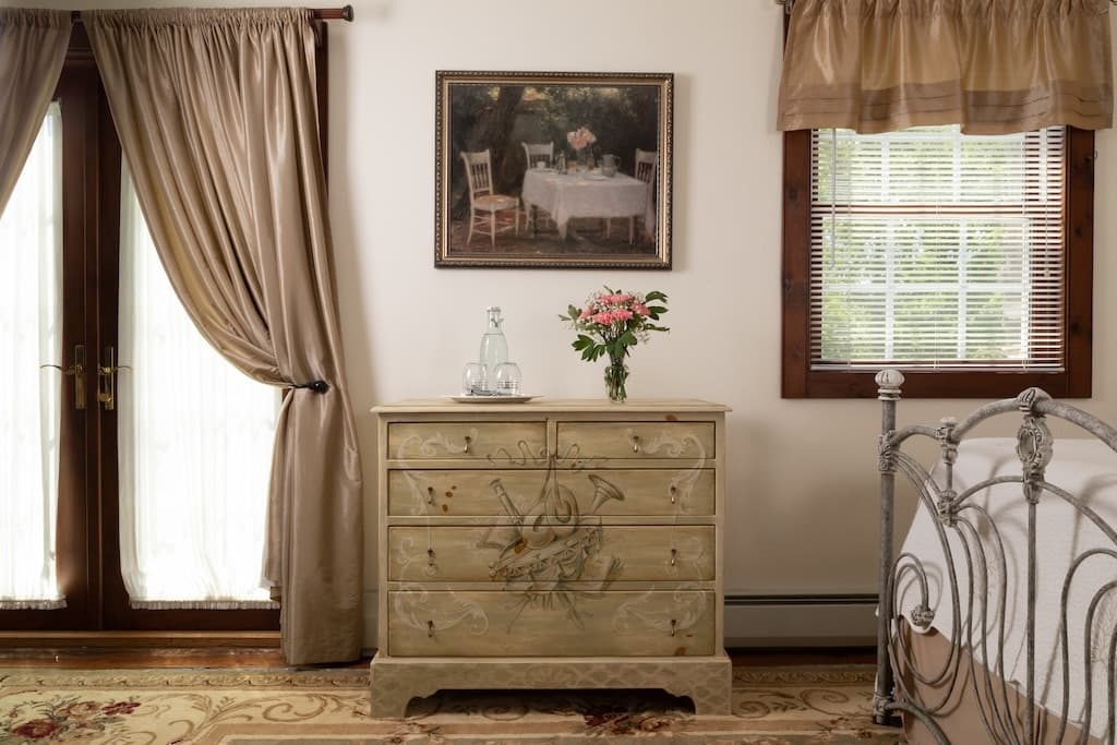 A cozy bedroom featuring a painted dresser, floral arrangement, and a scenic painting of a table set for two by a window.