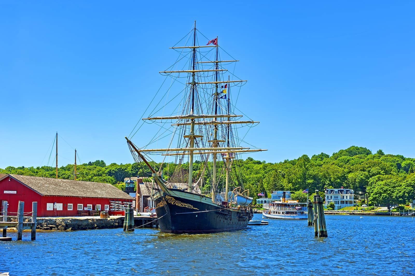 A tall ship anchored in a harbor near a red building and lush green trees under a clear blue sky.