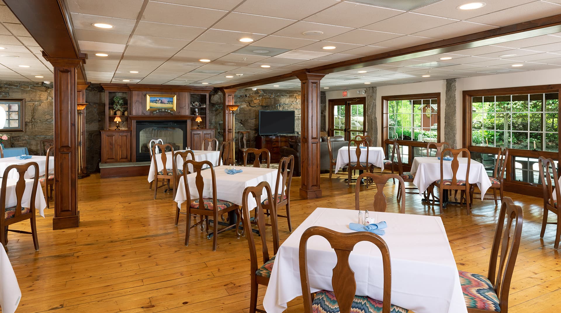A spacious dining room featuring wooden floors, tables with white tablecloths, and large windows.