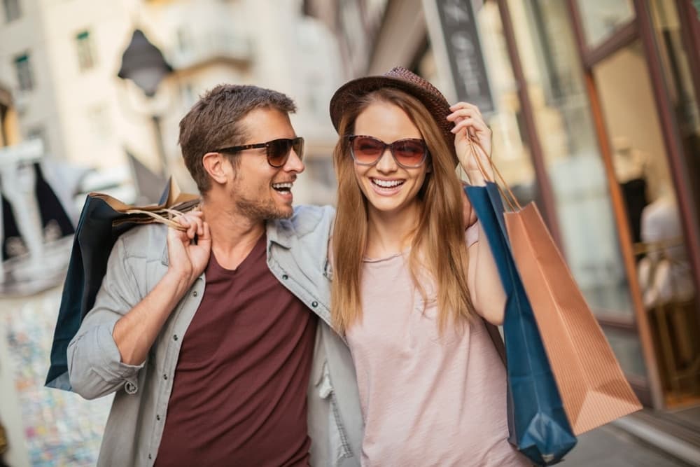 A smiling couple wearing sunglasses walks together, each holding shopping bags.