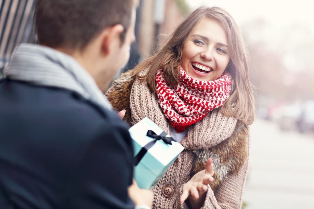 A woman joyfully reacts as a man hands her a gift on a chilly day.