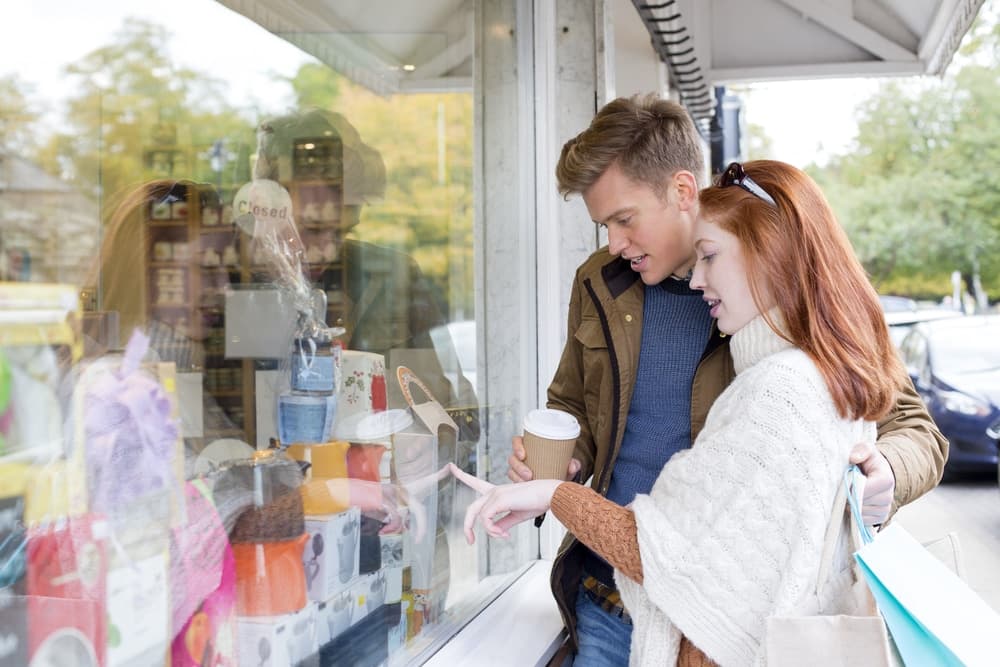 A couple gazes at shop items through a window while enjoying a coffee.