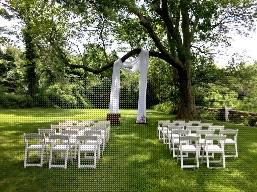 A wedding ceremony setup with white chairs arranged on a grassy lawn beneath a tree canopy.