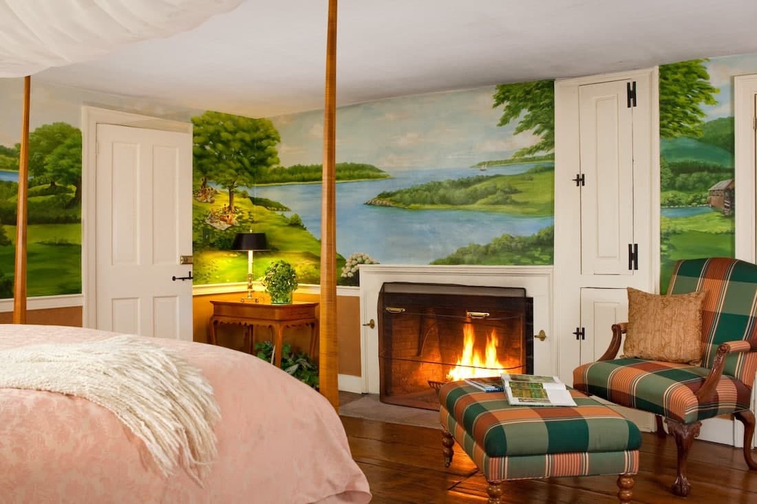 A cozy bedroom featuring a scenic mural, a four-poster bed, a fireplace, and a comfortable armchair.