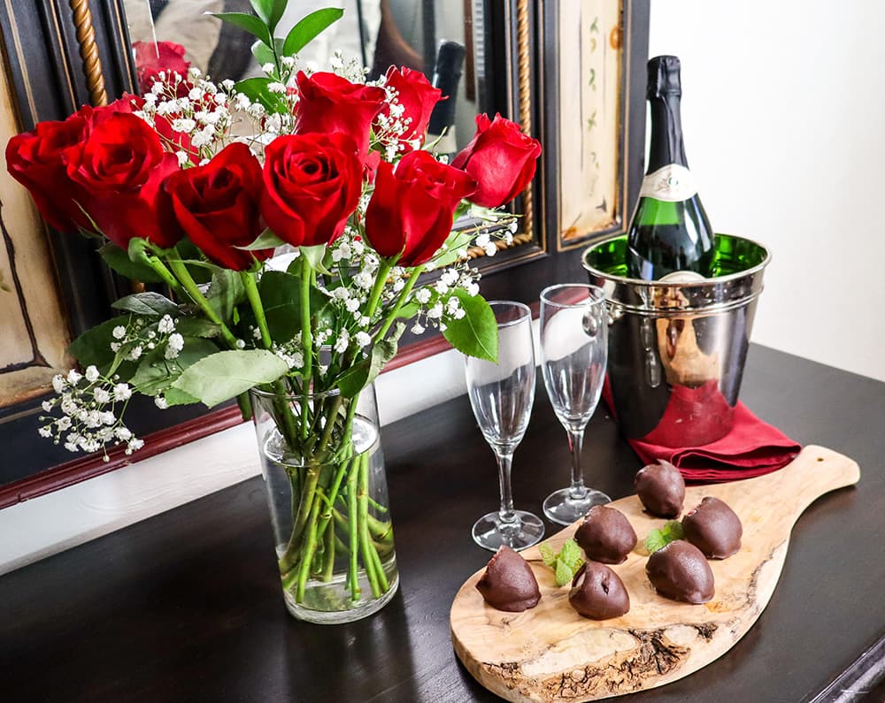 A vase of red roses sits next to champagne, flutes, and chocolate treats on a wooden platter.