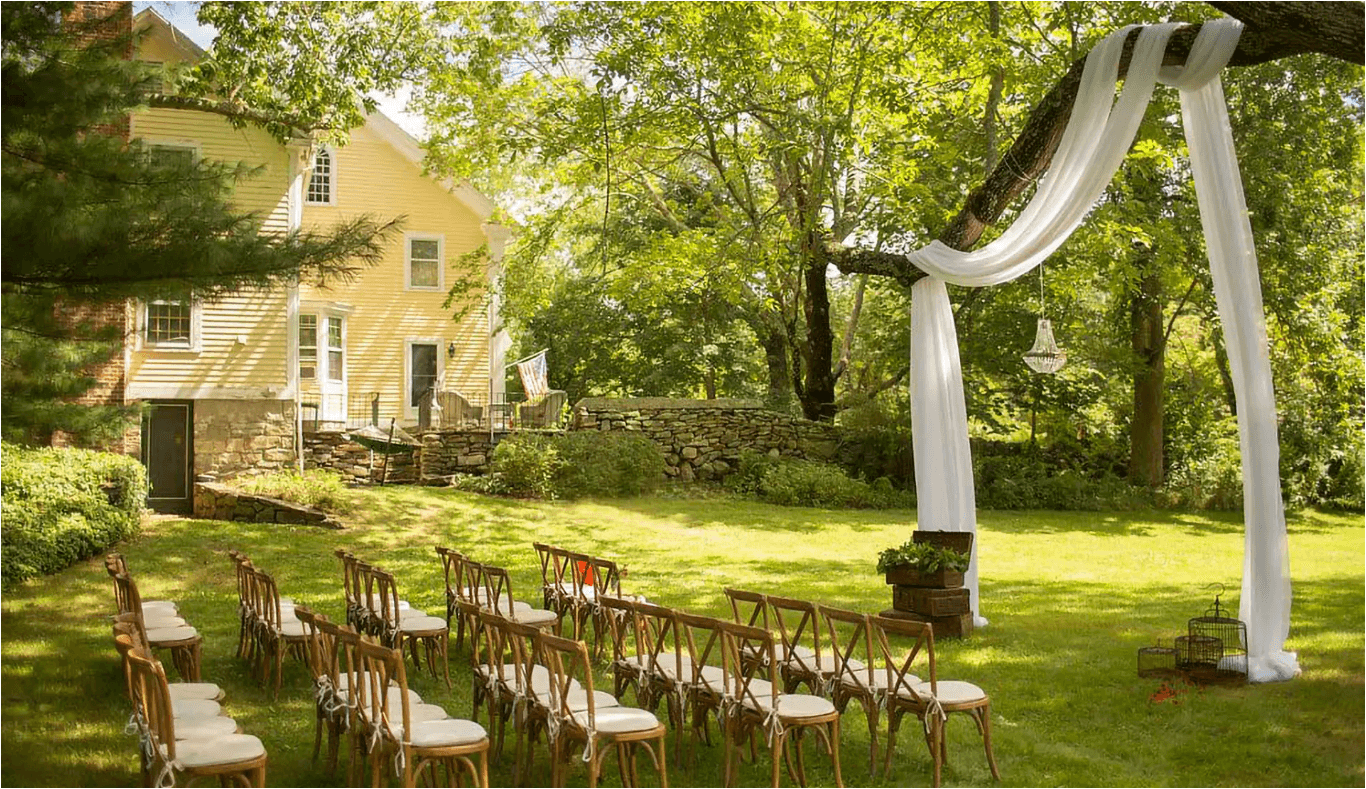 A charming outdoor wedding setup featuring rows of wooden chairs and a decorated archway beneath lush green trees by a historic yellow house.