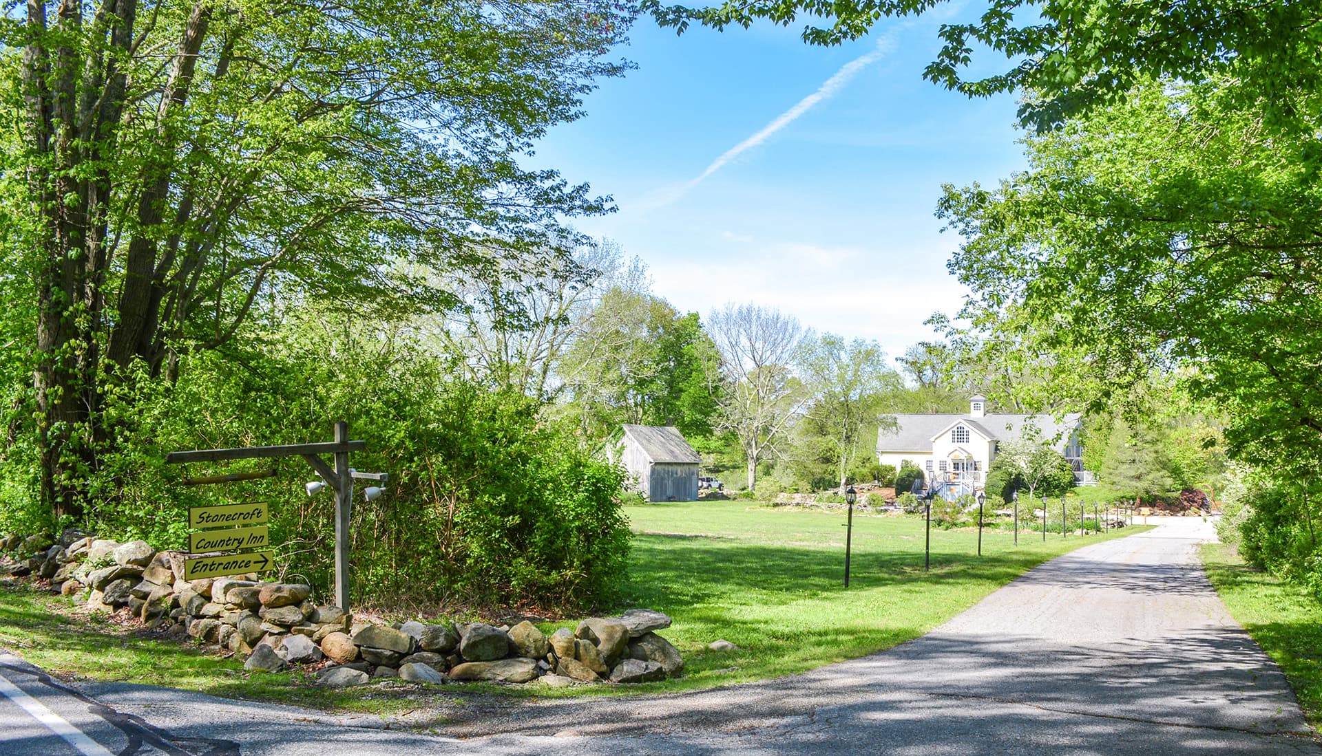 A gravel road leads to a country house surrounded by lush green trees and open fields.