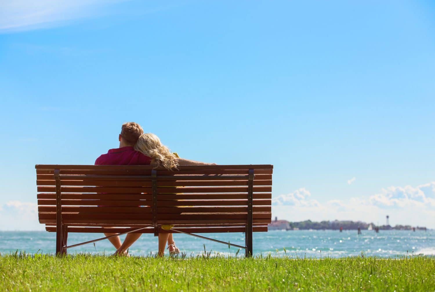 A couple sits together on a bench overlooking a serene water view under a clear blue sky.