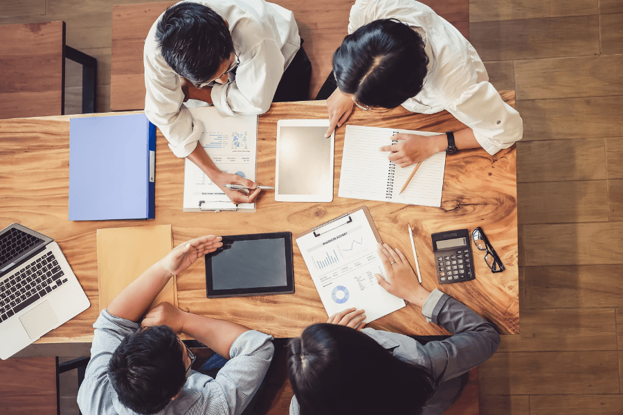 Aerial view of four people collaborating at a wooden table with laptops, documents, and a tablet.