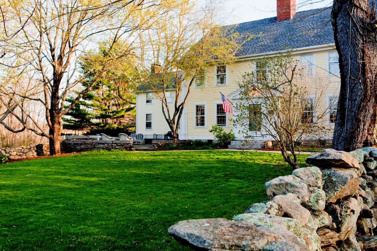A charming, light-colored house with an American flag, surrounded by green grass and trees.