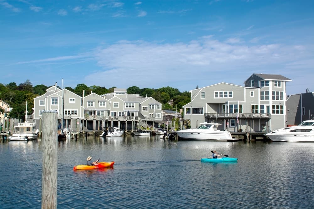 Two kayakers paddle in a calm harbor lined with modern gray houses and boats on a sunny day.