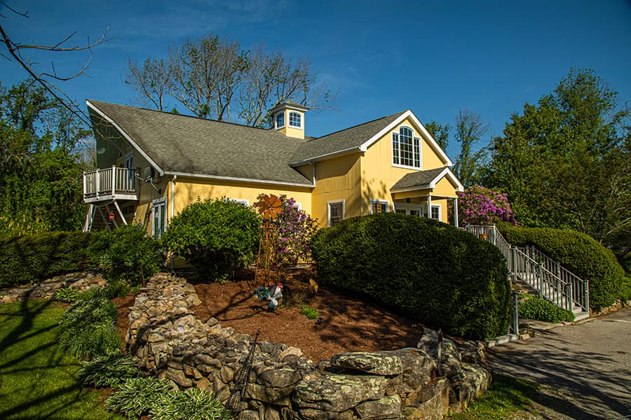 A yellow house surrounded by lush greenery and stone landscaping.
