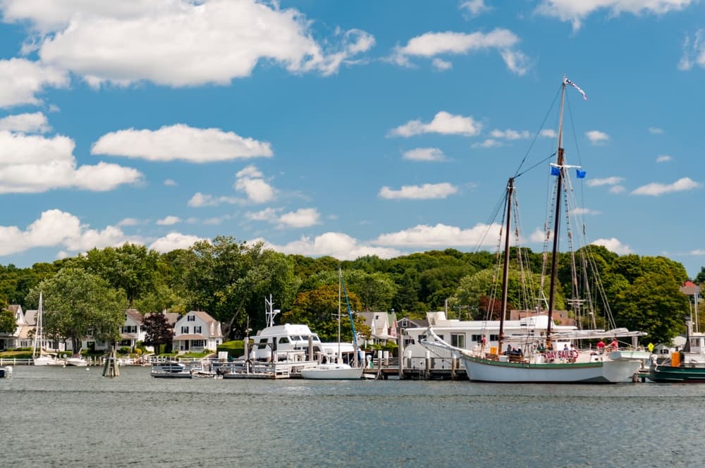 A peaceful harbor scene featuring sailboats docked beside lush greenery and quaint houses under a bright blue sky with fluffy clouds.