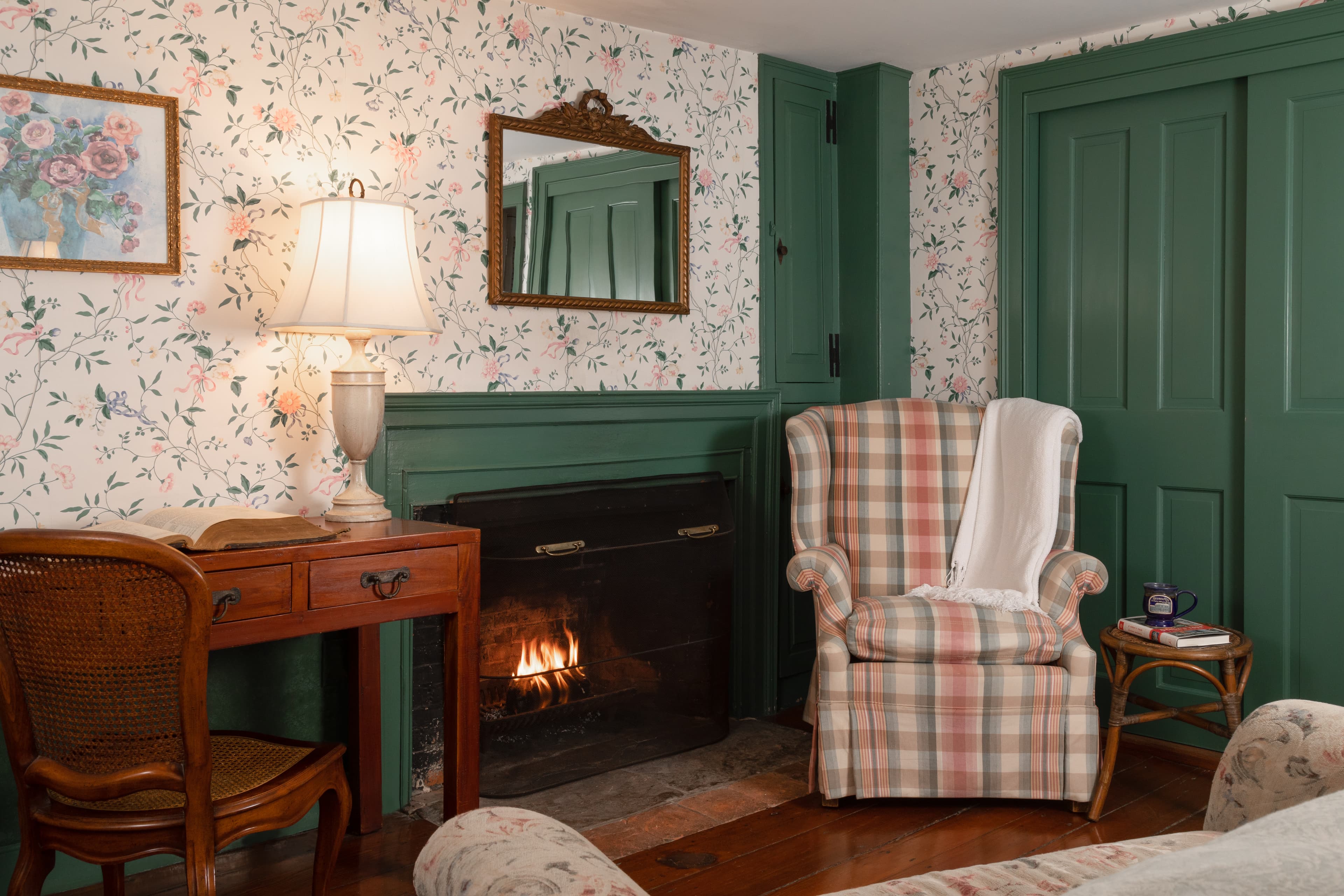 A cozy room featuring a plaid armchair, a small desk, a fireplace, and floral wallpaper.