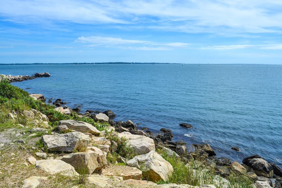 A rocky shoreline meets a calm blue sea under a clear sky.