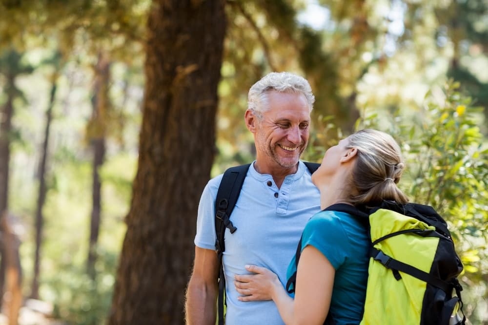 A man and woman smile at each other while standing in a forest, both wearing hiking gear.