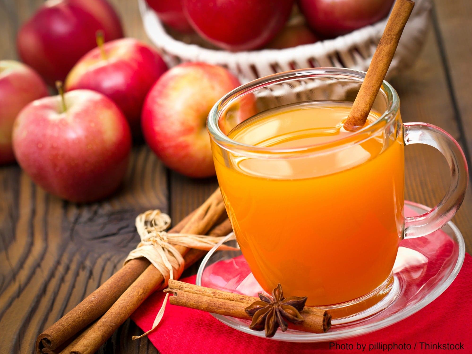 A glass of spiced apple cider with cinnamon sticks sits on a plate next to fresh apples.
