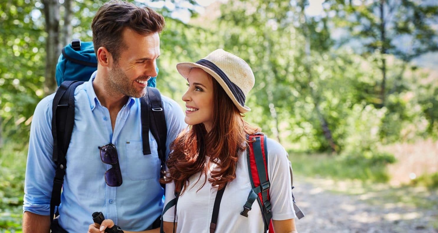 A smiling couple with backpacks shares a joyful moment while hiking on a forest trail.