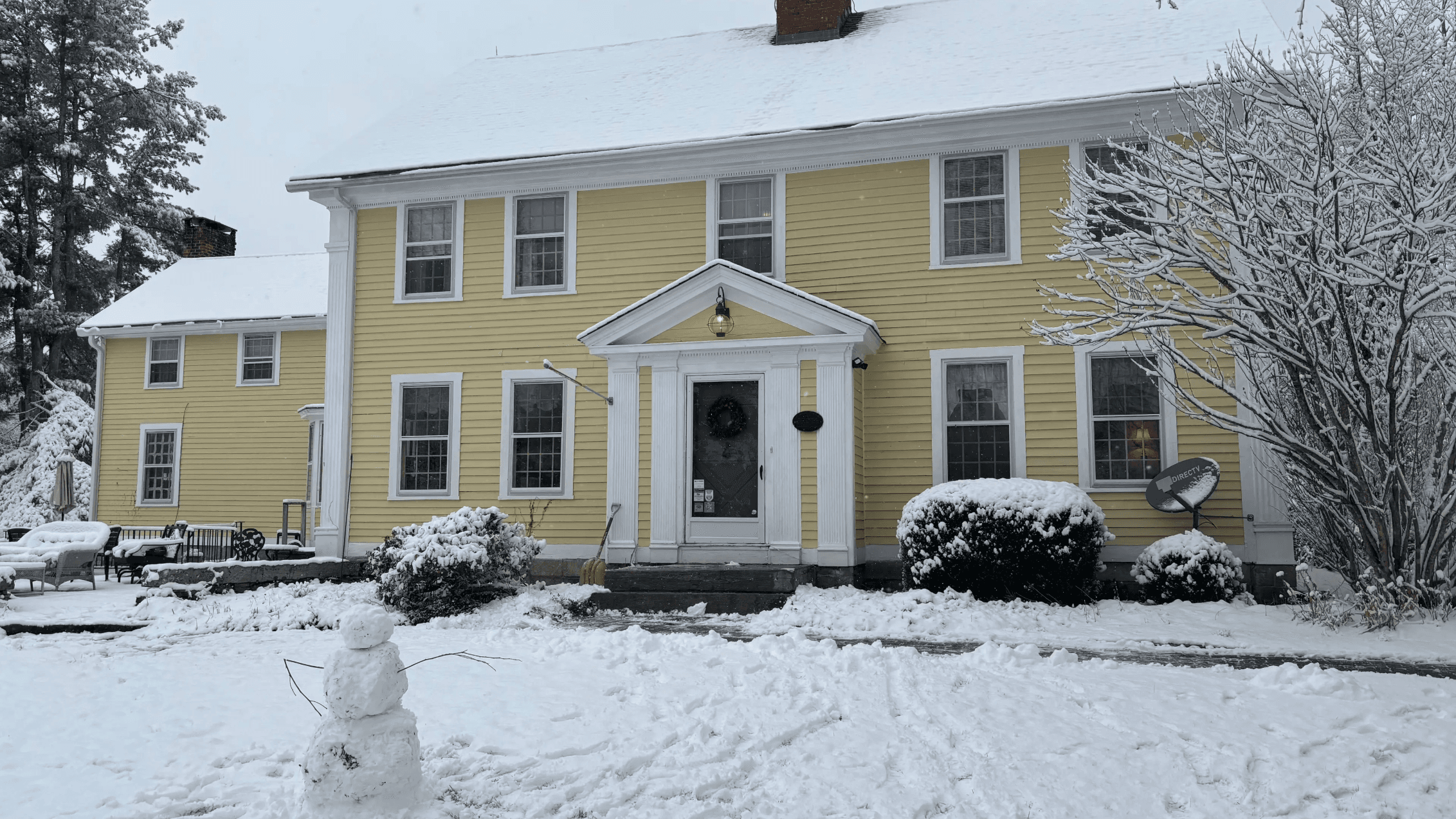 A yellow two-story house with snow-covered surroundings and a small snowman in the foreground. A yellow two-story house with snow-covered surroundings and a small snowman in the foreground.