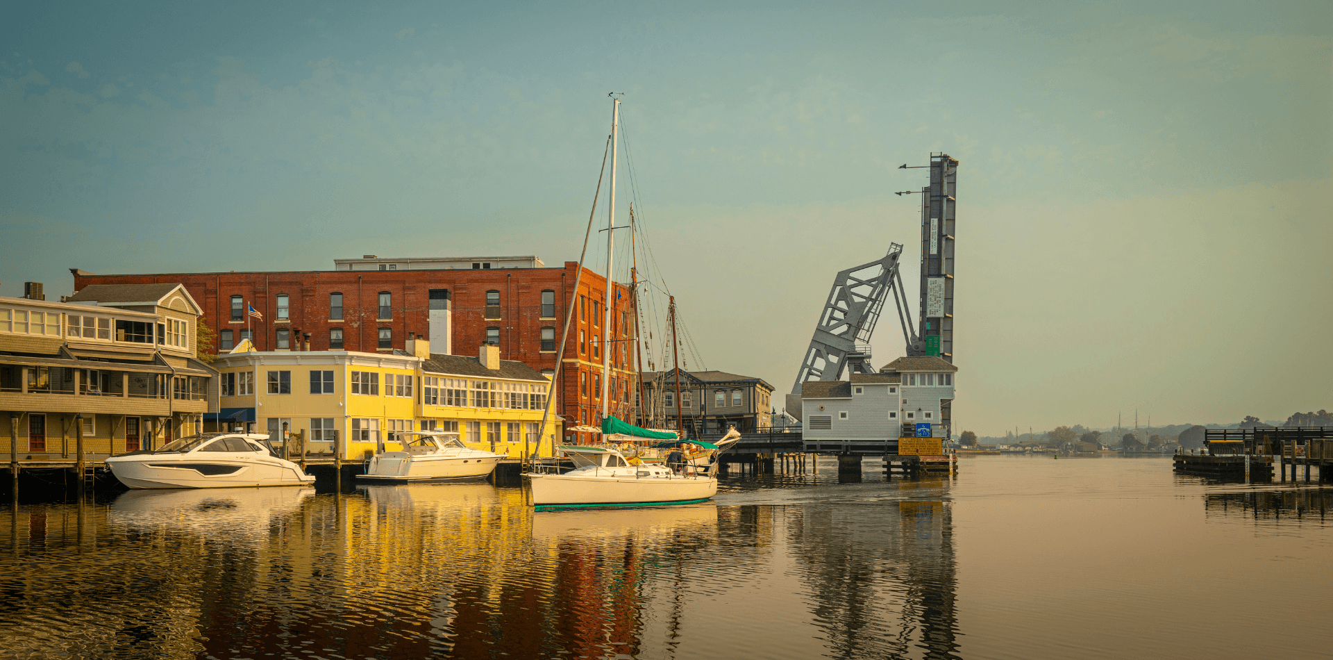 A marina scene featuring boats docked alongside historic buildings and a lift bridge reflecting in calm waters.