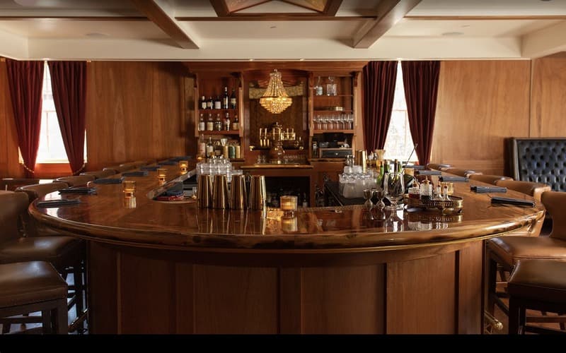 A stylish wooden bar with leather seating, dim lighting, and a chandelier, featuring various bottles and glassware.