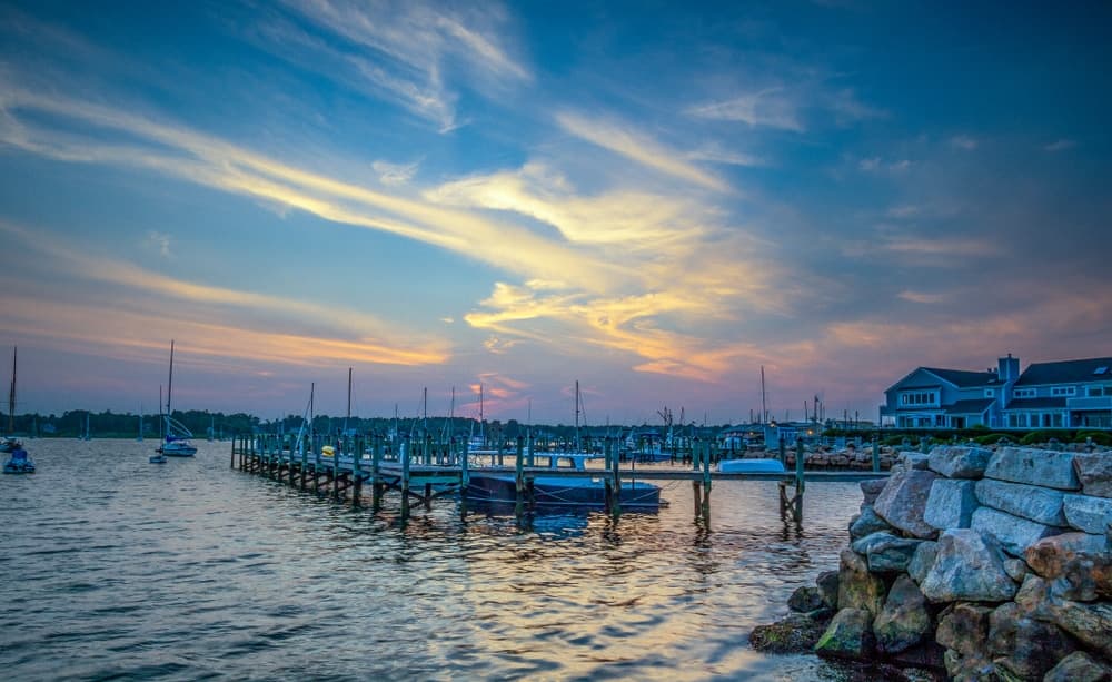 A tranquil sunset over a marina with sailboats and a wooden dock beside stone-banked water.