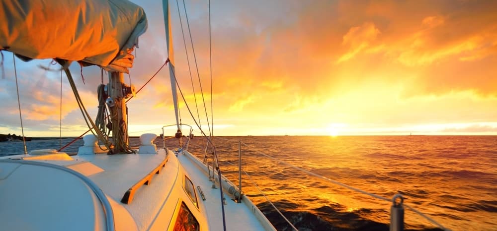 A sailboat glides through water as the sun sets, illuminating the sky in vibrant hues.