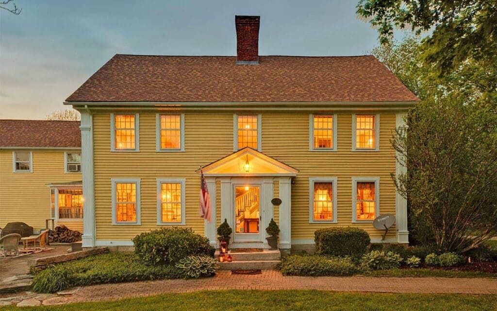 A large yellow colonial house with warm lights in the windows and a front porch surrounded by greenery.