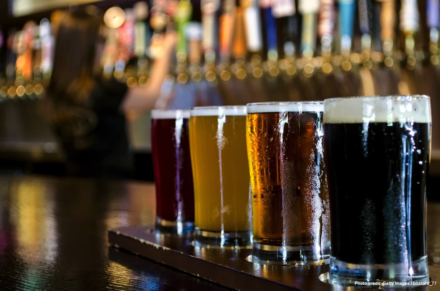 A flight of five colorful beer glasses on a bar counter with a blurred bartender in the background.