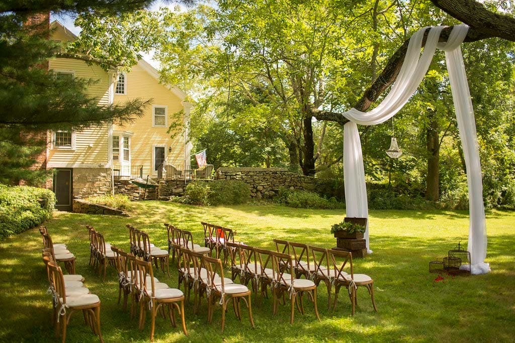 A picturesque outdoor wedding setup featuring rows of wooden chairs beneath a tree with white drapery.