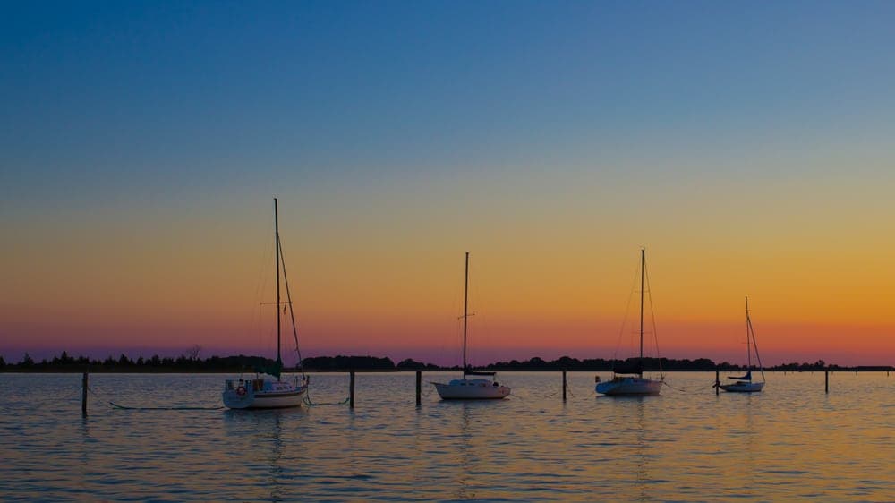 Five sailboats are moored at dusk, silhouetted against a vibrant sunset sky.