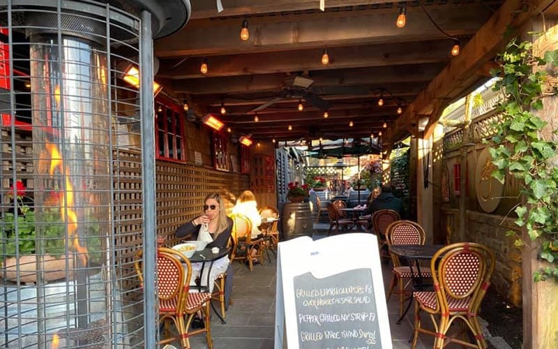 Outdoor dining area with string lights and a chalkboard menu, featuring patrons at tables under a wooden canopy.