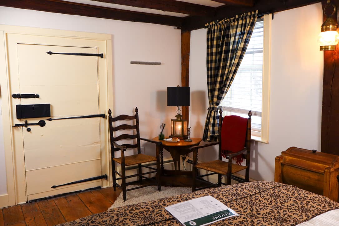 A warm cozy bedroom with dark wood exposed beams, a Dutch door and sitting area with two high back chairs with a table and lamp.