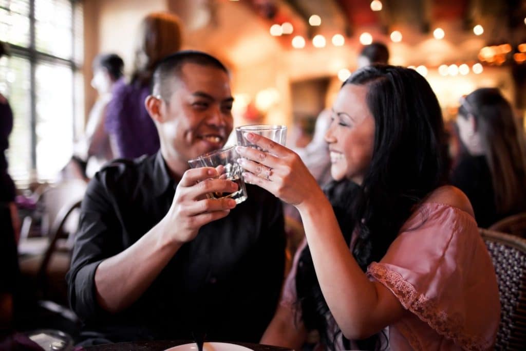 A couple smiles and clinks glasses together in a lively restaurant setting.