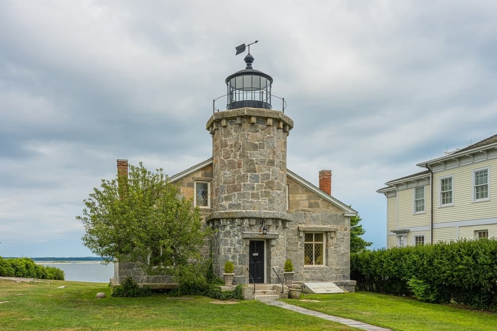 A stone lighthouse stands beside a grassy area and adjacent buildings under a cloudy sky.
