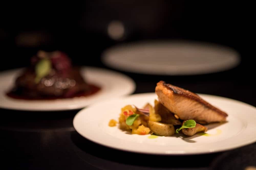 A plate with a piece of seared salmon and colorful vegetable medley is in focus, with another dish blurred in the background.
