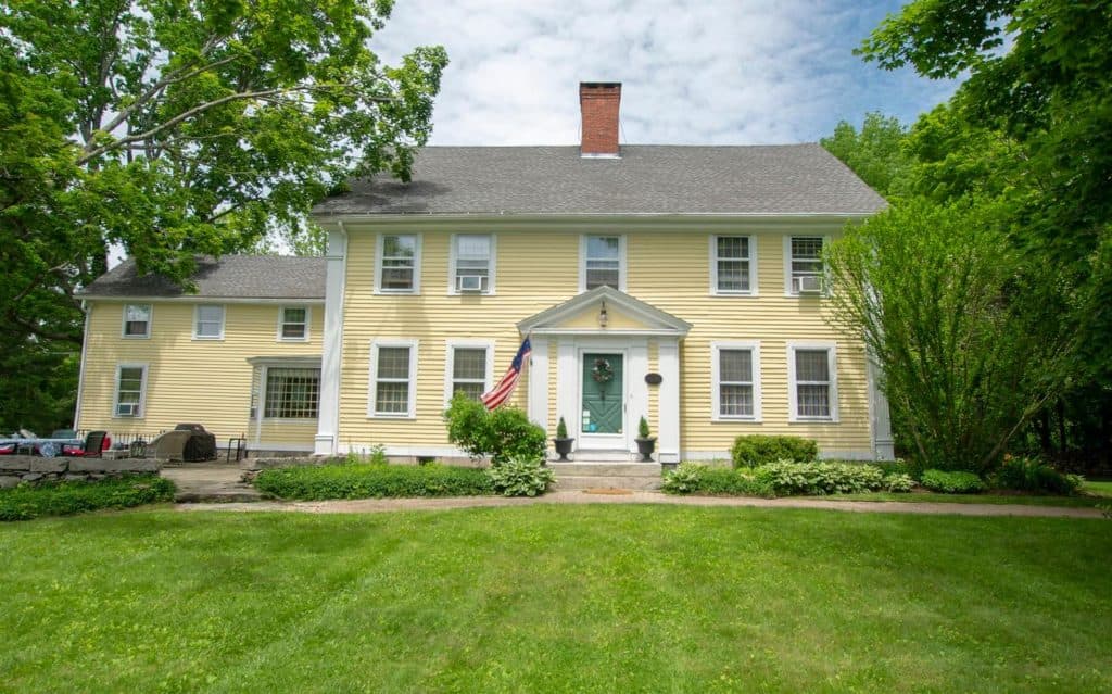 A large, yellow colonial-style house with a green door and American flag, surrounded by lush greenery and lawns under a partly cloudy sky.