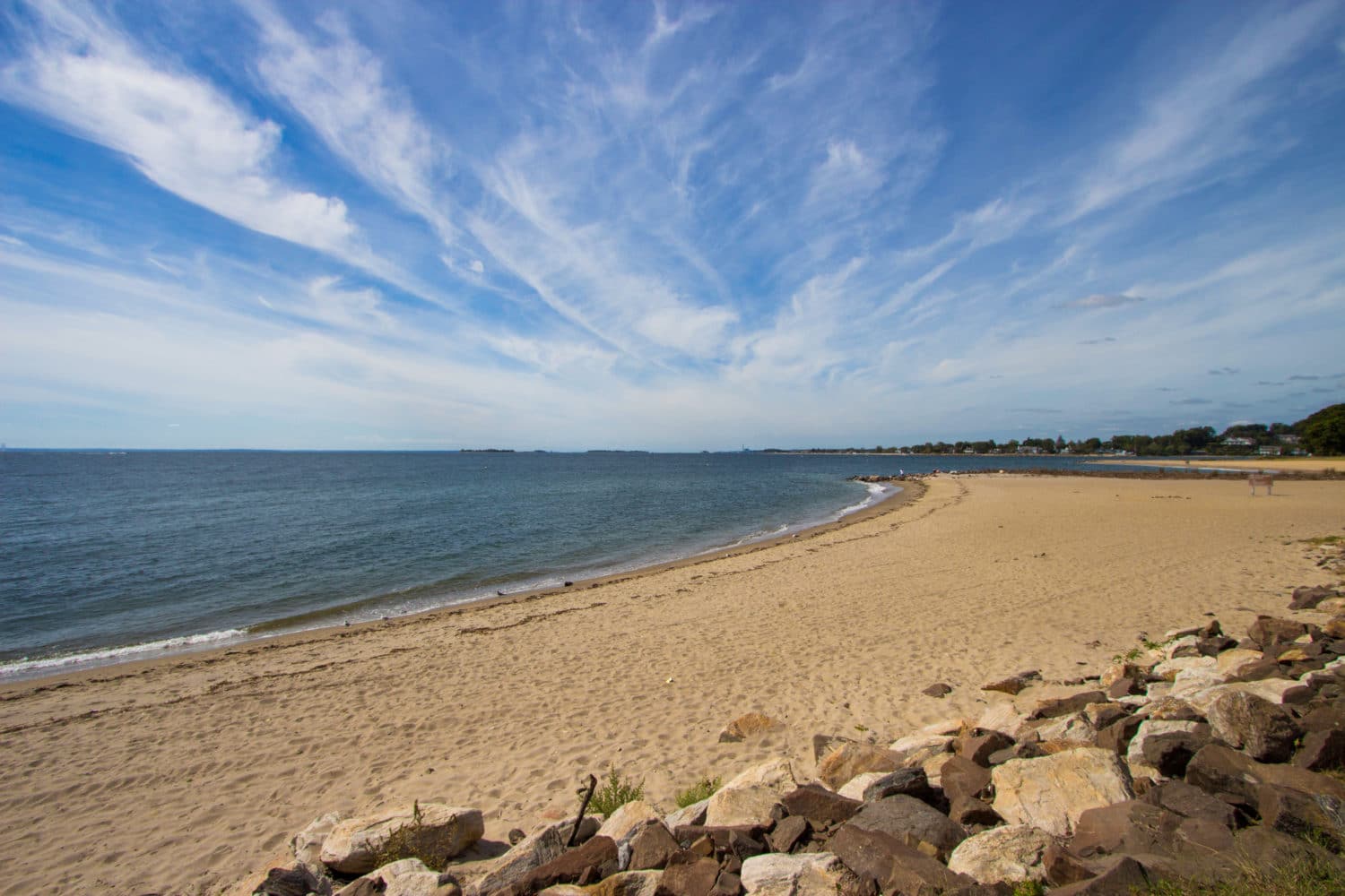 A sandy beach curves along a calm shoreline under a blue sky with wispy clouds.