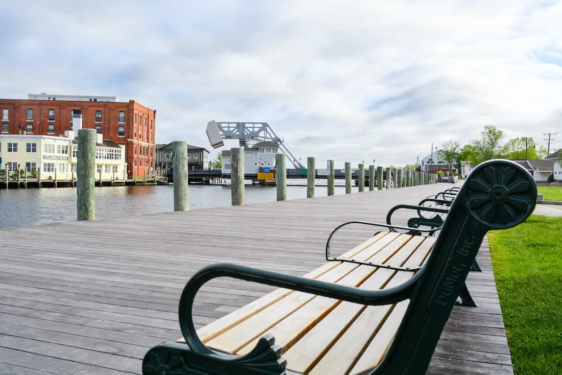 A wooden boardwalk lined with benches stretches alongside a calm waterway, with buildings and a drawbridge in the background.