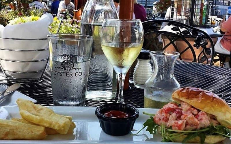 A plate of lobster roll with fries, accompanied by a glass of white wine and a water glass, set on a patio table.