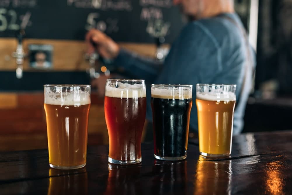 Four glasses of assorted beer on a bar, with a person in the background pouring from a tap.