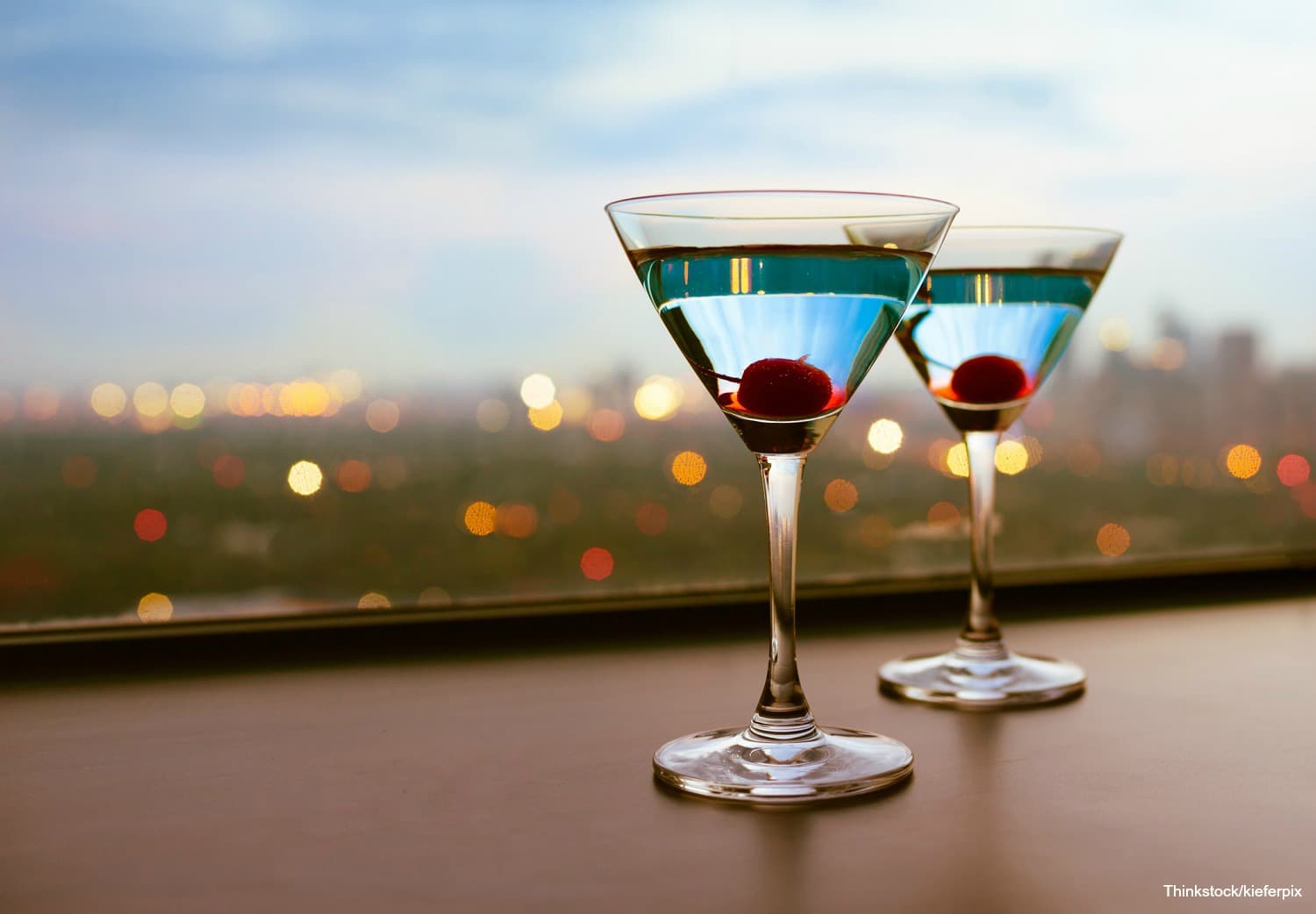 Two martini glasses with blue liquid and cherries sit against a blurred city skyline.