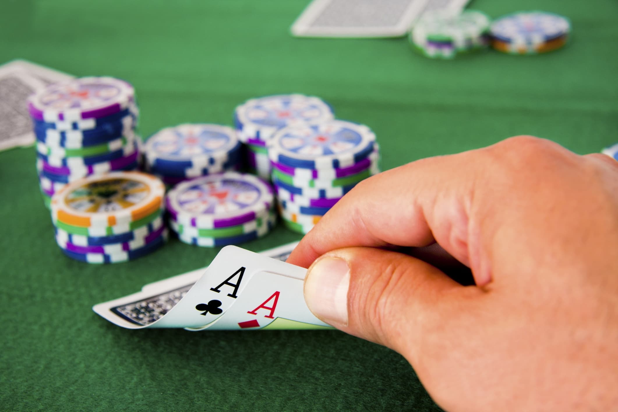 A hand holding a pair of aces over a green poker table with stacked chips in the background.