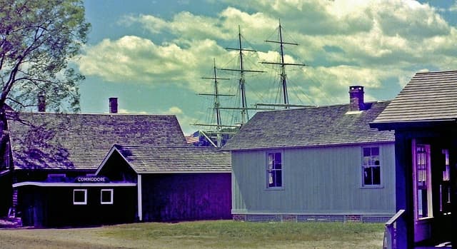 Historic buildings with a tall sailing ship in the background under a cloudy sky.