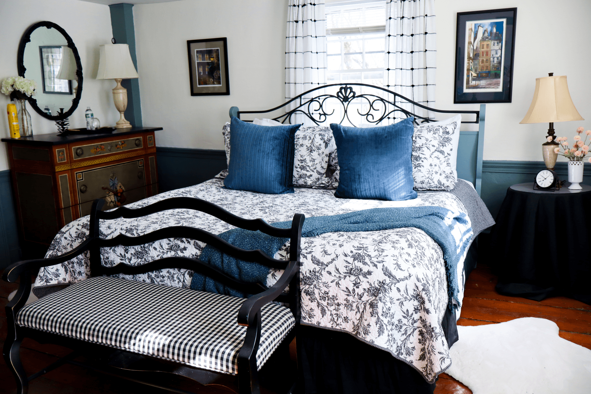 A cozy bedroom featuring a floral-patterned bedspread, two blue accent pillows, a wooden bench, and framed artwork on the walls.