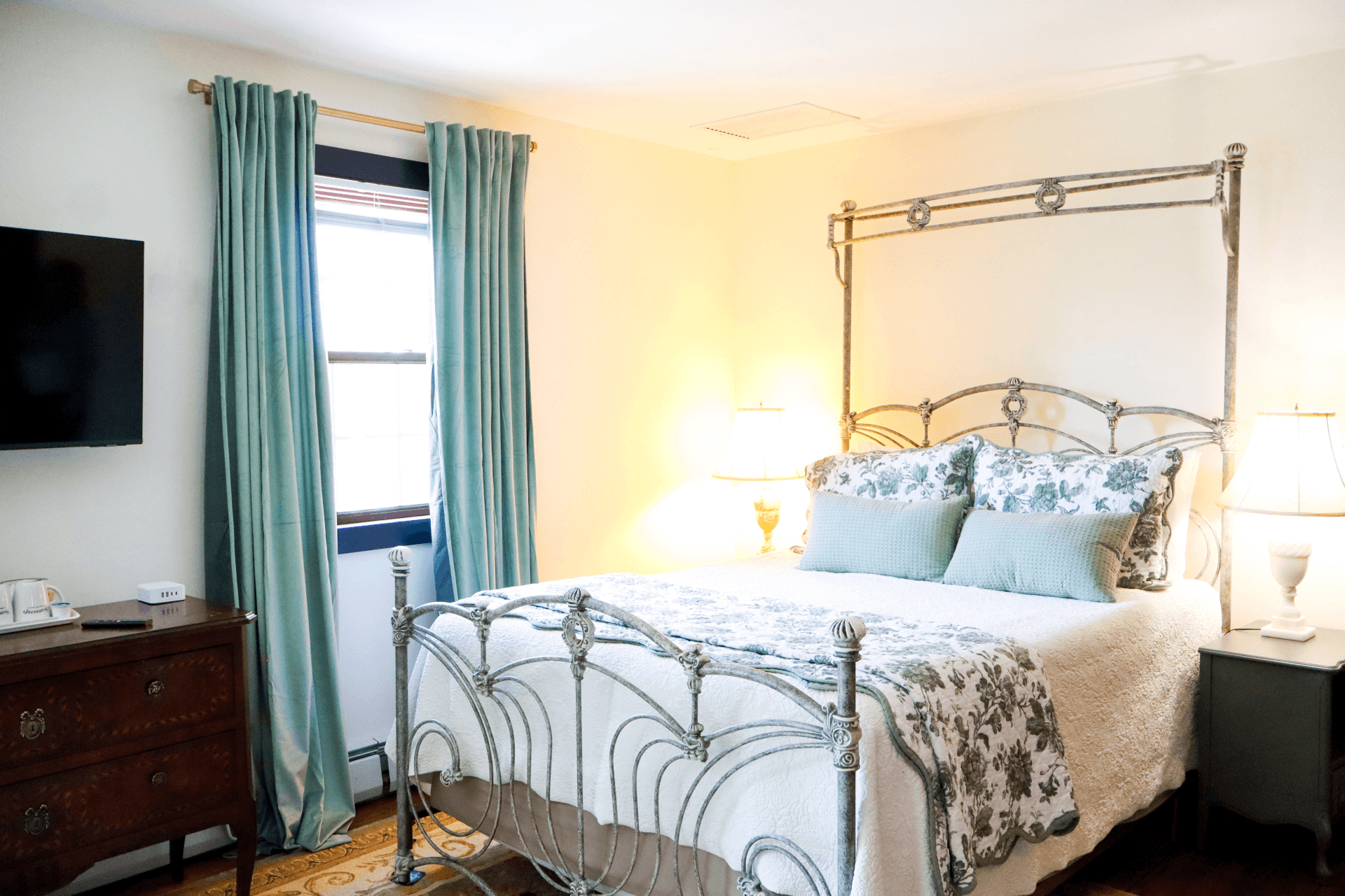 A light-filled bedroom featuring a metal bed frame, floral bedding, a wooden dresser, and soft green curtains.