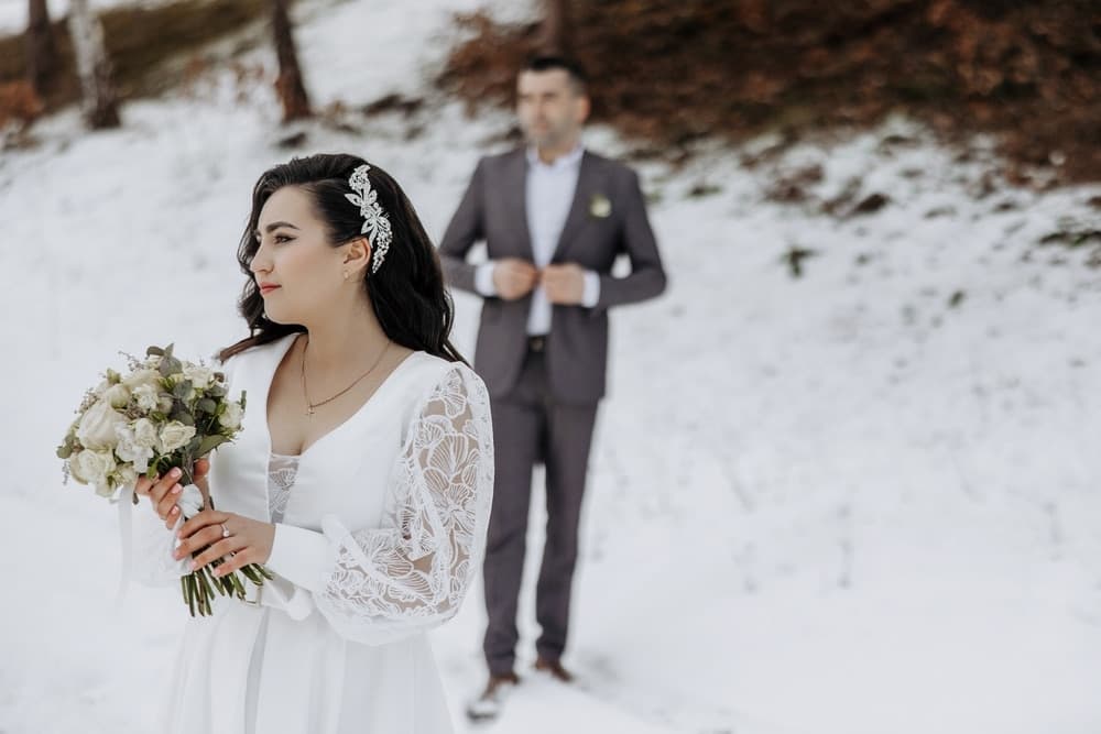 A bride holding a bouquet stands in the snow, while a groom adjusts his jacket in the background.