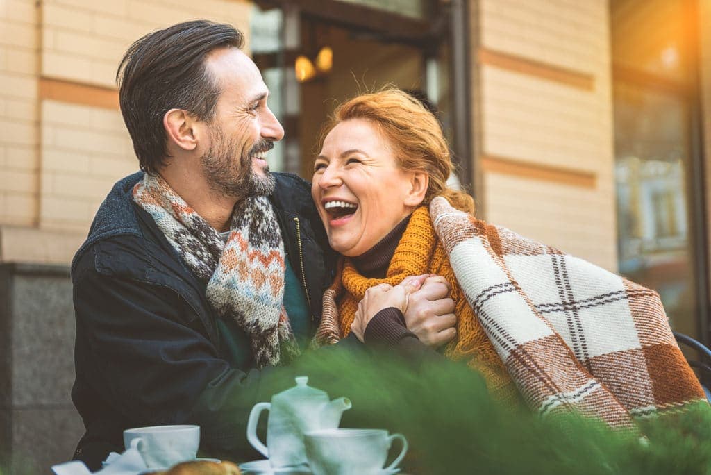 A couple in warm scarves joyfully laughing together at a café table.