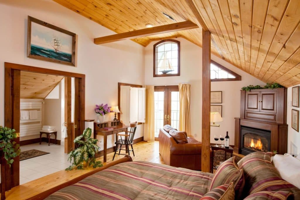 A cozy bedroom with wooden beams, a fireplace, and a seating area near large windows.